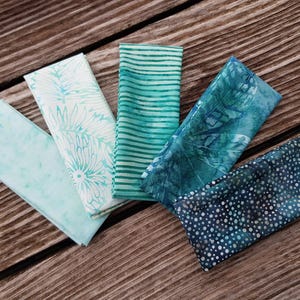 Può includere: Cinque tovaglioli di stoffa piegati in tonalità di blu e verde sono disposti su una superficie di legno. I tovaglioli presentano vari motivi, tra cui righe, disegni floreali e pois. I colori vanno dal turchese chiaro al verde acqua scuro.