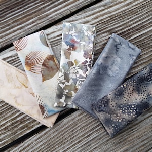 Puede incluir: Cinco servilletas de tela plegadas con varios diseños, incluyendo diseños botánicos y abstractos. Los colores varían del beige y marrón al azul y gris. Las servilletas están dispuestas sobre una superficie de madera envejecida.