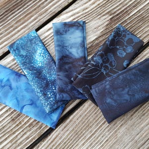 Può includere: Un assortimento di quadrati di tessuto piegati in tonalità di blu, disposti su una superficie di legno. I tessuti presentano vari motivi, tra cui disegni maculati, effetti acquerello e stampe floreali. La tavolozza dei colori va dal chiaro all'indaco scuro.