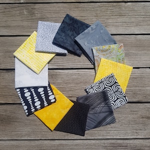 Könnte beinhalten: Eine Sammlung von 12 Stoffquadraten in verschiedenen Mustern in Schwarz, Weiß und Gelb. Die Quadrate sind in einem Kreis auf einer Holzoberfläche angeordnet.