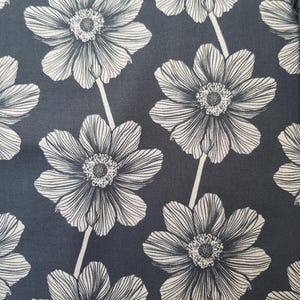May include: Close-up of a fabric with a repeating floral pattern. Large, stylized white flowers with detailed petals are set against a dark gray background. The flowers are connected by thin, white stems, creating a visually appealing textile design.