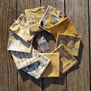 Könnte beinhalten: Eine Sammlung von zehn Stoffquadraten in verschiedenen Gelb- und Cremetönen. Die Stoffquadrate zeigen Designs wie Schmetterlinge, Blumen, Karten und eine Bingokarte. Die Stoffquadrate sind in einem Kreis auf einer Holzoberfläche angeordnet. Der Text "Eclectic Elements" ist auf einem Etikett in der Mitte des Kreises gedruckt.