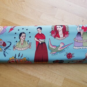 May include: Rolled fabric featuring a light blue background with Frida Kahlo-inspired tattoo-style illustrations. The design includes portraits, floral elements, a swallow, and text in Spanish. The fabric is suitable for various craft projects.