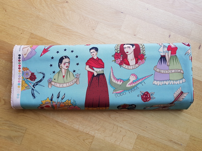 May include: Rolled fabric featuring a light blue background with Frida Kahlo-inspired tattoo-style illustrations. The design includes portraits, floral elements, a swallow, and text in Spanish. The fabric is suitable for various craft projects.
