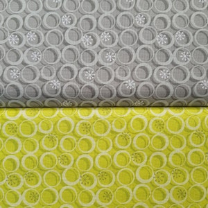 Może przedstawiać: Dwa próbki tkaniny z powtarzającym się wzorem nakładających się kół i płatków śniegu. Górna próbka jest szara z białymi kołami i płatkami śniegu. Dolna próbka jest żółta z białymi kołami i płatkami śniegu.