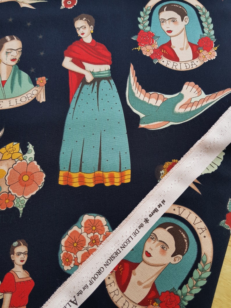Op de afbeelding: Een donkerblauwe stof met een herhalend patroon van Frida Kahlo-afbeeldingen, bloemen en een duif. De stof heeft de tekst "FRIDA" en "VIVA."