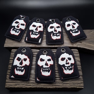 Könnte beinhalten: Acht schwarze Stoff-Schlüsselanhänger mit weißen Totenkopf-Designs und roten Umrissen. Die Schlüsselanhänger sind in zwei Reihen zu vieren auf einer Holzoberfläche angeordnet.