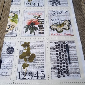 Op de afbeelding: Een rol vintage-stof met een botanisch en numeriek ontwerp. De cr&egrave;mekleurige stof heeft verschillende rechthoekige panelen met bloemenillustraties, zaadzakjes en tekst. Een zwarte pen ligt op de stof.