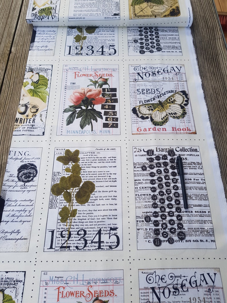 Op de afbeelding: Een rol vintage-stof met een botanisch en numeriek ontwerp. De cr&egrave;mekleurige stof heeft verschillende rechthoekige panelen met bloemenillustraties, zaadzakjes en tekst. Een zwarte pen ligt op de stof.