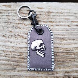 Könnte beinhalten: Ein grauer Stoffschlüsselanhänger mit einem schwarzen Totenkopfdesign und einem schwarzen Metallring und Clip.