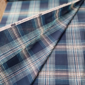 May include: A close-up of a navy blue fabric with a white and teal plaid pattern. The fabric is folded in half and the label reads "DYKE (LIGHT)".
