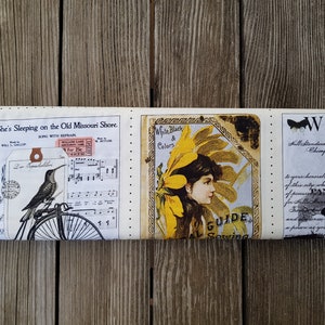 Op de afbeelding: Een opgerolde stof met vintage-stijl illustraties en tekst. Het ontwerp bevat een kolibrie op een fiets, bladmuziek en een vrouwenportret met de tekst "White, Black & Colors". De stof is cr&egrave;mekleurig met zwarte, gele en bruine accenten.