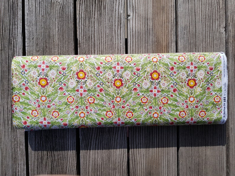 May include: A white fabric with a green, red, and yellow floral pattern. The fabric has a repeating pattern of flowers and leaves. The fabric is on a wooden surface.