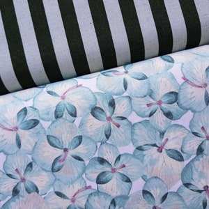 Może przedstawiać: Biała tkanina z powtarzającym się wzorem niebieskich kwiatów akwarelowych. Tkanina jest złożona na czarno-białej tkaninie w paski.