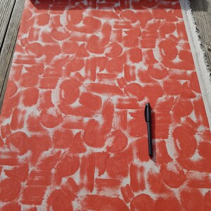 May include: Red fabric with a white abstract brushstroke pattern. The fabric is folded and a black pen is lying on top of it.