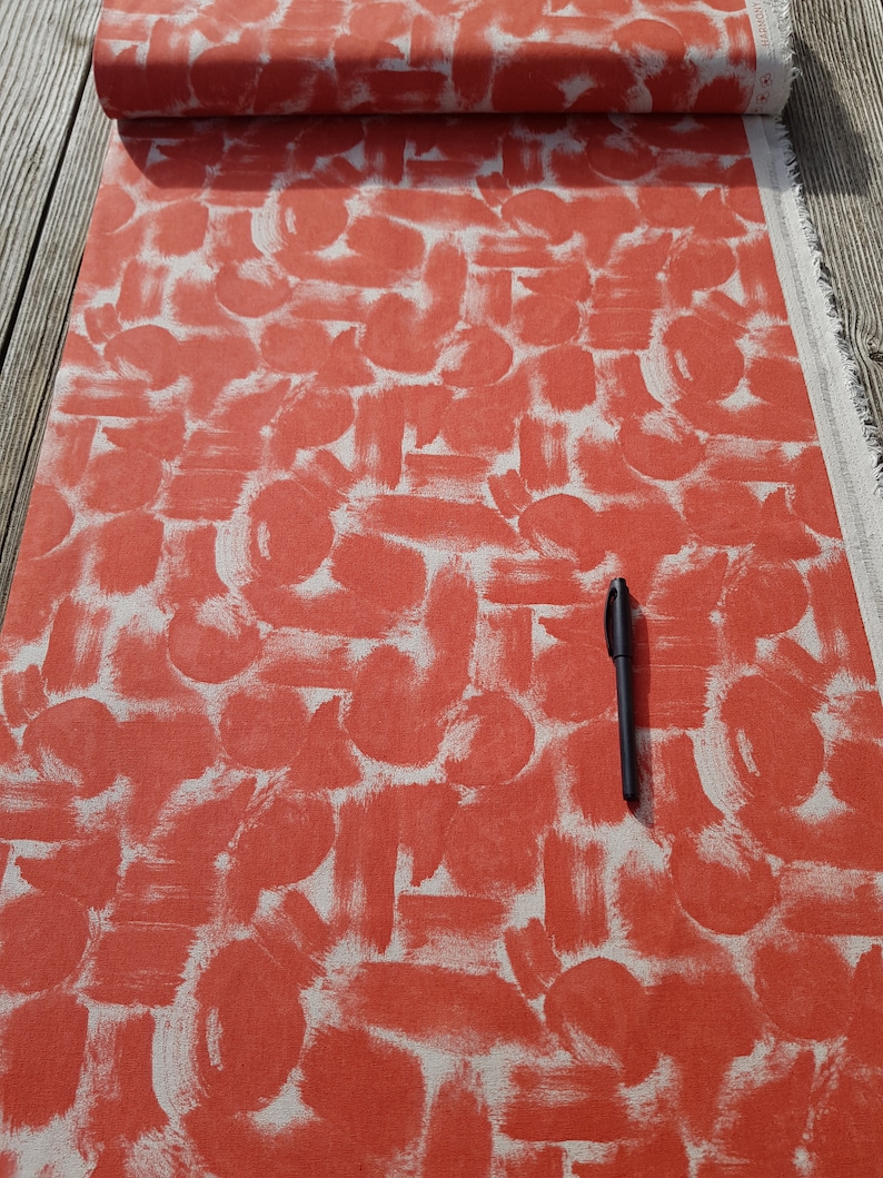 May include: Red fabric with a white abstract brushstroke pattern. The fabric is folded and a black pen is lying on top of it.