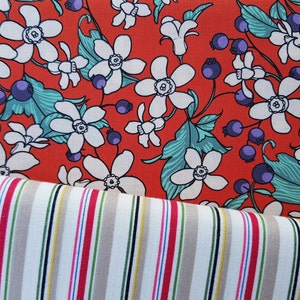 May include: Red fabric with a white floral pattern and green leaves. The fabric is layered over a white and beige striped fabric.