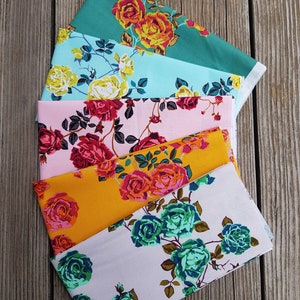 May include: Six fabric swatches with floral patterns featuring roses in various colors, including pink, red, orange, yellow, teal, and green. The fabric swatches are arranged in a fan shape on a wooden surface.