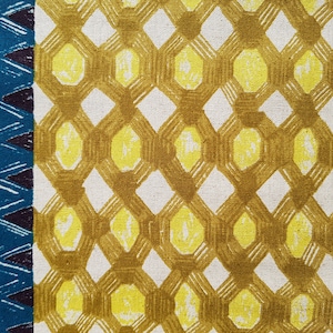 Op de afbeelding: Een witte stof met een herhalend patroon van gele en bruine geometrische vormen. Het patroon bestaat uit overlappende zeshoeken en ruiten.