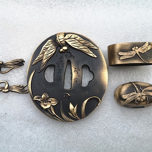 Op de afbeelding: Een set van vier gouden metalen Japanse zwaardbeslag, of tsuba, met libellenontwerpen. De belangrijkste tsuba is rond en heeft een zwarte achtergrond met gouden accenten. De andere drie tsuba zijn kleiner en hebben een eenvoudiger ontwerp.