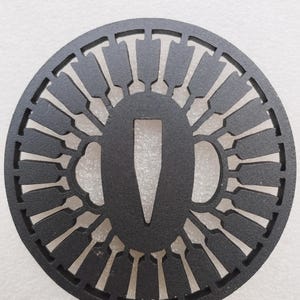 May include: A black, circular tsuba, a Japanese sword guard, with an intricate openwork design. The guard features a central opening for the sword tang and a series of radiating, stylized shapes around the perimeter. The tsuba is made of metal.