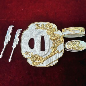 Op de afbeelding: Een zilveren en gouden Japanse zwaardknop, of tsuba, met een bloemmotief. De tsuba is een decoratief stuk dat wordt gebruikt om de hand te beschermen tegen het blad. De tsuba is ook een kunstwerk en wordt vaak versierd met ingewikkelde ontwerpen.