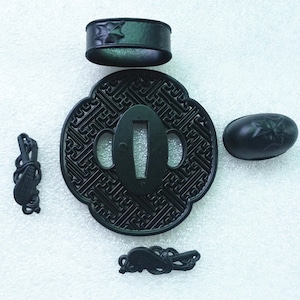 Op de afbeelding: Een set zwarte metalen zwaardbeslag, inclusief een tsuba (handbescherming), fuchi (kraag) en twee kashira (knopkappen). De tsuba heeft een geometrisch patroon, terwijl de fuchi en kashira bloemmotieven hebben.