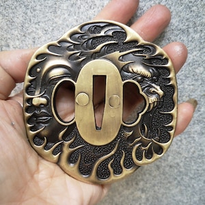 Op de afbeelding: Een messing tsuba, een decoratieve zwaardbeschermer, met een gedetailleerd ontwerp van een draak en golven. De tsuba is een traditioneel Japans zwaardaccessoire.