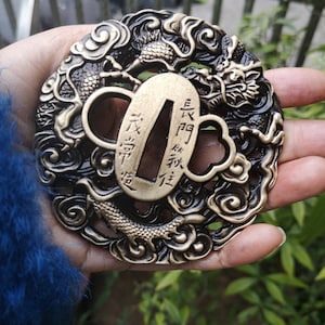 Op de afbeelding: Een ronde, zwart-gouden metalen tsuba, een decoratieve zwaardbeschermer, met een gedetailleerd drakenontwerp en ingewikkelde wolkenpatronen. De tsuba heeft een rechthoekige opening voor het zwaardblad en bevat Japanse karakters.