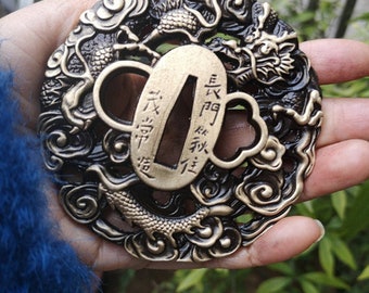 May include: A circular, black and gold metal tsuba, a decorative sword guard, with a detailed dragon design and intricate cloud patterns. The tsuba has a rectangular opening for the sword blade and features Japanese characters.
