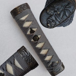 May include: Two gray and white sword handles with intricate metal details. The handles are wrapped in a gray cord and have a white diamond pattern. The metal details are embossed with a design.