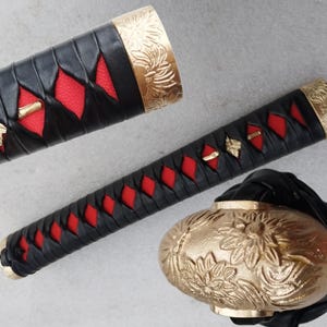 May include: Close-up of a katana handle with a black leather wrap and red diamond-shaped accents. The handle features gold-colored floral designs on the pommel and collar. The overall design is ornate and detailed.