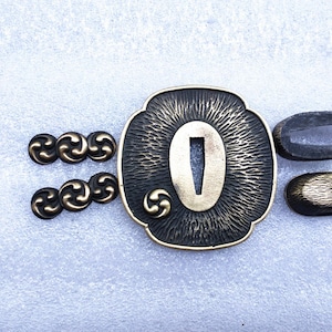 Op de afbeelding: Een set van vier gouden metalen zwaardbeslag, waaronder een tsuba (handbescherming) met een getextureerd oppervlak en een ronde opening, twee kleine decoratieve stukken en een zwart metalen stuk met een rechthoekige opening.