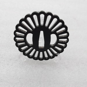 Op de afbeelding: Zwarte metalen tsuba, een decoratieve bescherming voor een Japans zwaard, met een ronde vorm en een zonnestralenontwerp. De tsuba heeft een centrale opening voor het blad.