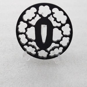 Op de afbeelding: Zwarte metalen tsuba, een decoratieve zwaardbeschermer, met een ronde vorm en een ontwerp van herhalende wolkvormen. De tsuba heeft een centrale opening voor het zwaardblad.