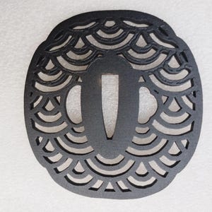Op de afbeelding: Een zwarte, ovaalvormige tsuba, een Japanse zwaardbeschermer, met een decoratief golfpatroon. De tsuba heeft een centrale opening voor de zwaardtang en is gemaakt van een donker, mogelijk metalen materiaal. Het ontwerp is ingewikkeld.