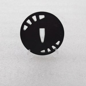 Puede incluir: Un tsuba circular negro, una guarda decorativa para una espada japonesa, con una abertura central y una serie de aberturas más pequeñas alrededor del borde.