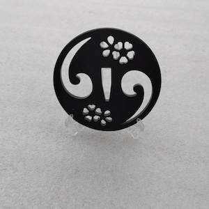 Op de afbeelding: Een zwarte ronde tsuba, een decoratieve bescherming voor een Japans zwaard, met een gestileerd bloemmotief en een centrale opening voor de zwaardtang.
