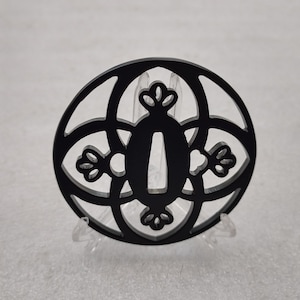 Op de afbeelding: Zwarte metalen tsuba, een decoratieve bescherming voor een Japans zwaard, met een cirkelvormig ontwerp met ingewikkelde geometrische patronen en een centrale opening.
