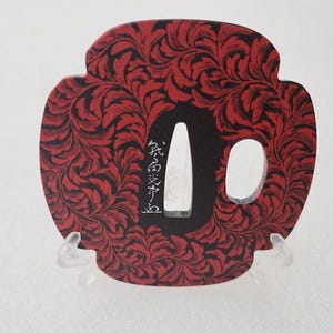 Puede incluir: Objeto decorativo rojo y negro con un patrón, con una abertura central y una abertura lateral más pequeña. El diseño presenta un patrón floral repetitivo. Se pueden ver caracteres japoneses en el objeto.