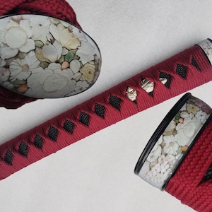 Op de afbeelding: Detail van een katana-handvat en schede. Het handvat is gewikkeld in rode stof met zwarte ruitvormige accenten. De schede en handvateinden hebben bloemmotieven in wit, geel en groen.