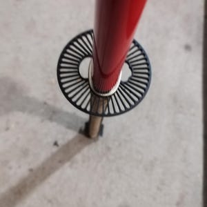 May include: A red metal pole with a black, circular, decorative element. The element has a radial design. The pole is attached to a small, wooden base. The image is taken from a high angle.