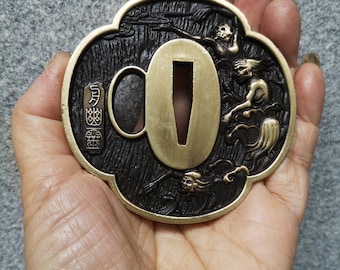 May include: A brass sword guard, or tsuba, with a black patina and intricate carvings of mythical creatures. The tsuba has a circular opening for the sword blade and a rectangular opening for the handle. The tsuba is decorated with a dragon and other mythical creatures.