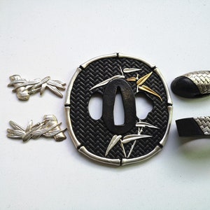 Puede incluir: Un tsuba de espada samurái negro y plateado con un diseño de bambú. El tsuba es una guarda decorativa que se utiliza para proteger la mano de la hoja. El tsuba está hecho de metal y tiene un fondo negro con hojas de bambú plateadas y un tallo de bambú dorado. El tsuba es una forma de arte japonesa tradicional y se utiliza a menudo como elemento decorativo en las espadas samuráis.