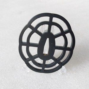 Op de afbeelding: Zwarte ronde tsuba, een decoratieve bescherming voor een Japans zwaard, met een geometrisch ontwerp van concentrische cirkels en stralende lijnen.