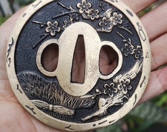 May include: A circular, brass tsuba, a decorative sword guard, with a black background. The tsuba features a stylised design of a bird in flight with cherry blossoms and a branch. The tsuba has a circular opening in the centre.