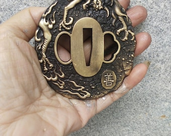 May include: A round, brass tsuba, a decorative sword guard, with a detailed design of two mythical creatures. The tsuba has a central opening for the sword blade and a raised inscription in Japanese characters.