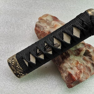 Peut inclure: Une poignée de katana noire et blanche avec des accents décoratifs en laiton. La poignée est enveloppée d'un cordon noir avec des panneaux blancs en forme de losange. Les extrémités de la poignée ont des capuchons en laiton ornés. La poignée repose sur une pierre rugueuse brun-rouge.