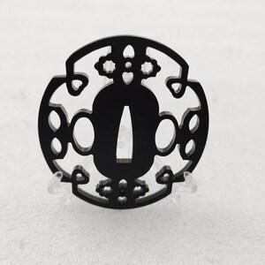 Op de afbeelding: Een zwarte Japanse katana tsuba, een zwaardbeschermer, met een ingewikkeld open ontwerp. De tsuba is cirkelvormig met een centrale opening en decoratieve uitsparingen. Het wordt tentoongesteld op een heldere standaard tegen een witte achtergrond.