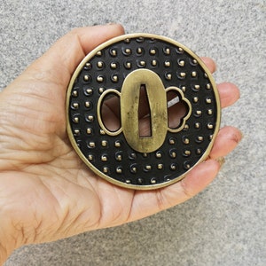 Peut inclure: Une garde d'épée ronde en métal noir et or avec un motif décoratif. La garde a une découpe pour la lame et deux plus petites découpes de chaque côté.